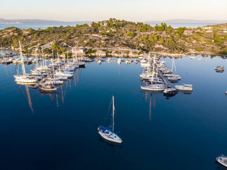 aegean-sea-port-with-multiple-moored-yachts-near-piers-greenery-blue-water-view-from-drone-greece
